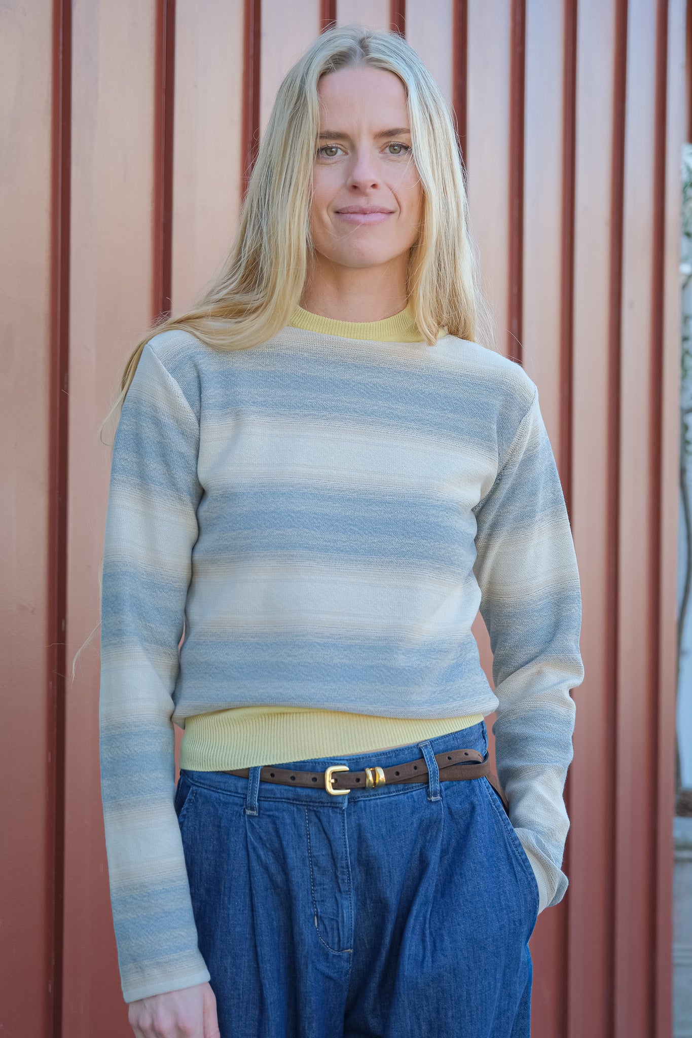 Woman wearing a striped sweater and blue jeans standing against a red wall.