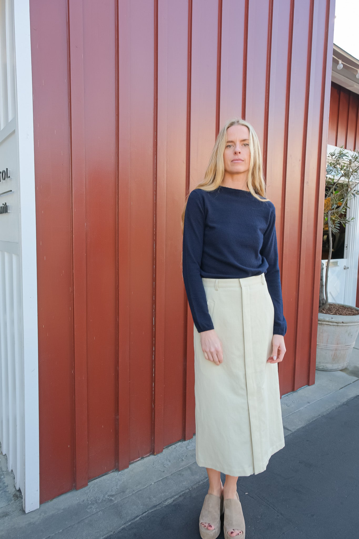 Woman standing in front of a red building wearing a navy top and beige skirt.