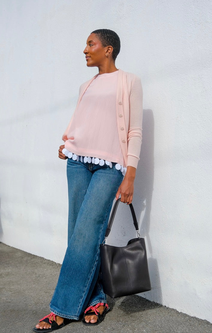 Woman wearing a light pink cardigan, blue jeans, and carrying a black handbag against a white wall.