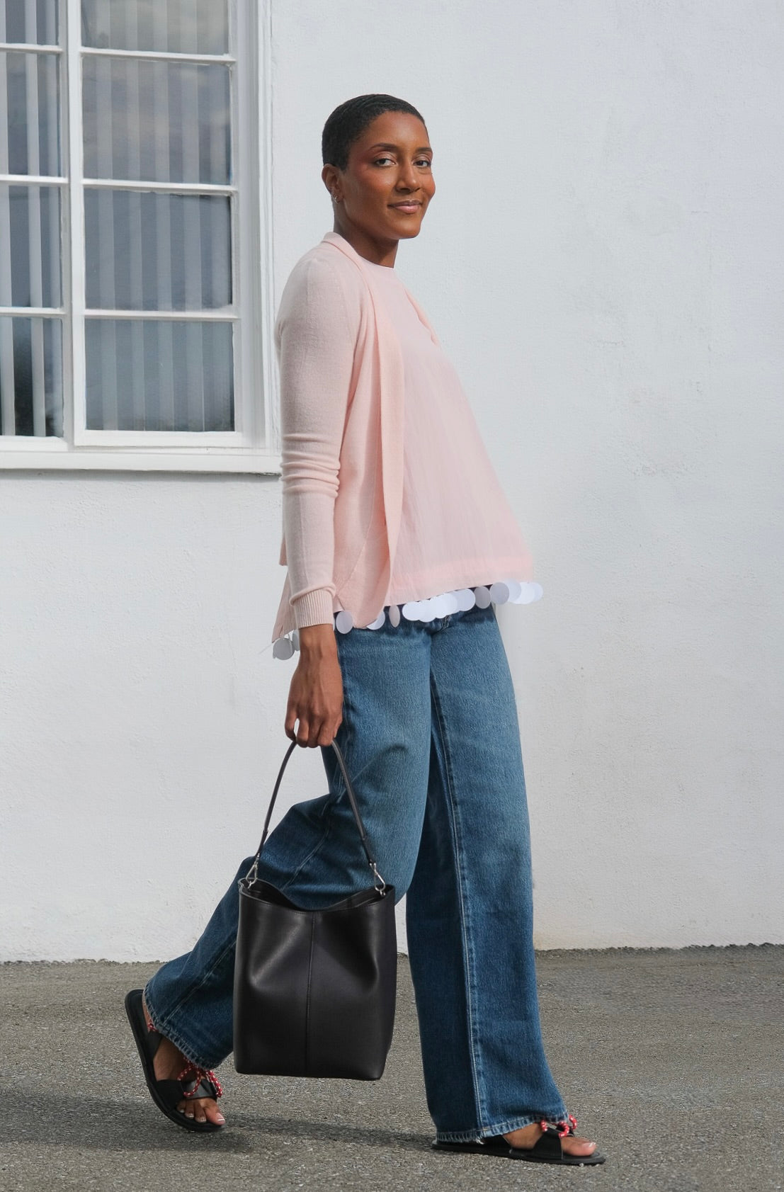 Woman wearing a light pink blouse and blue jeans, holding a black bag against a white wall.