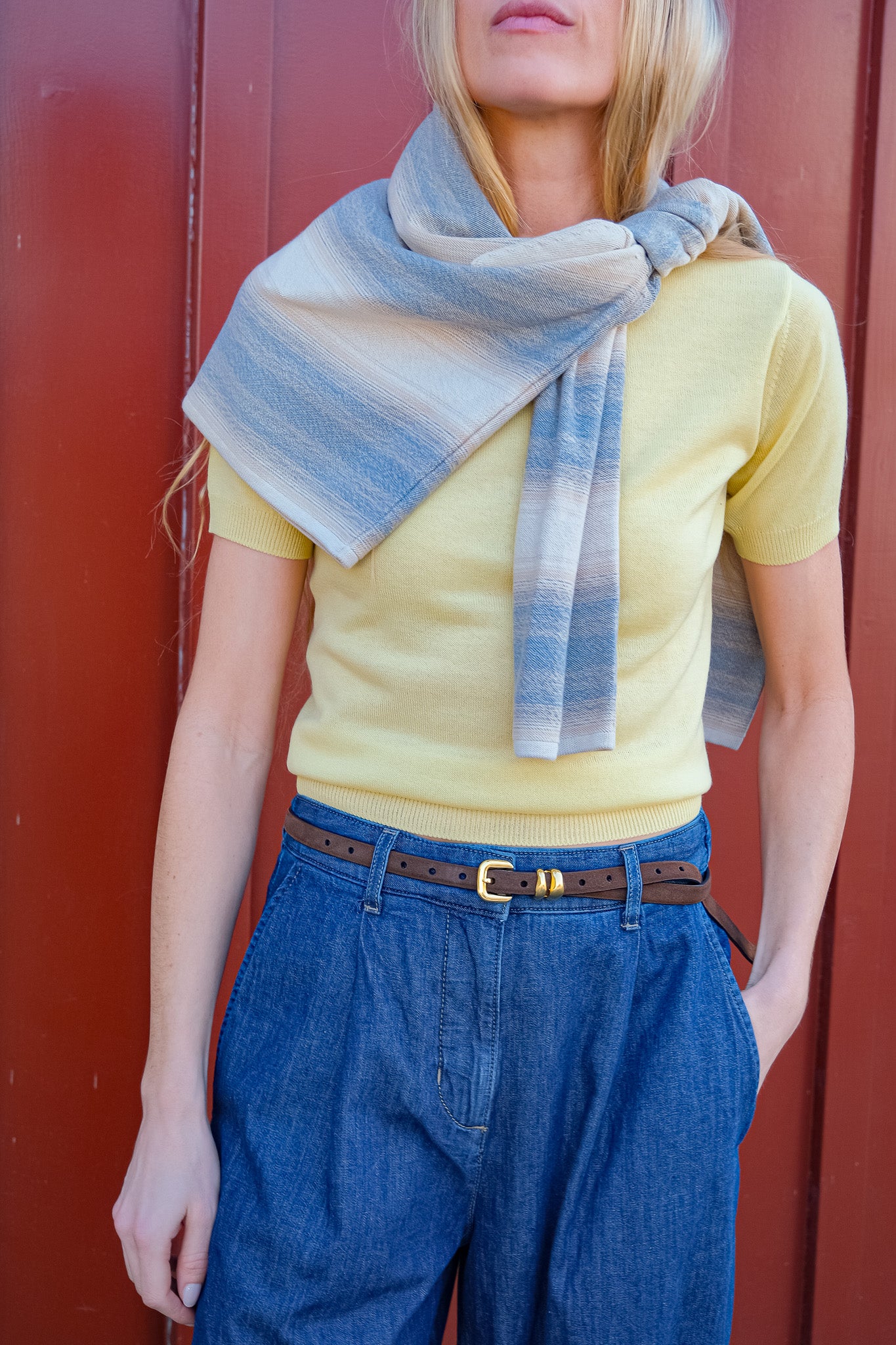 Person wearing a yellow top and blue jeans with a striped scarf, standing against a red background
