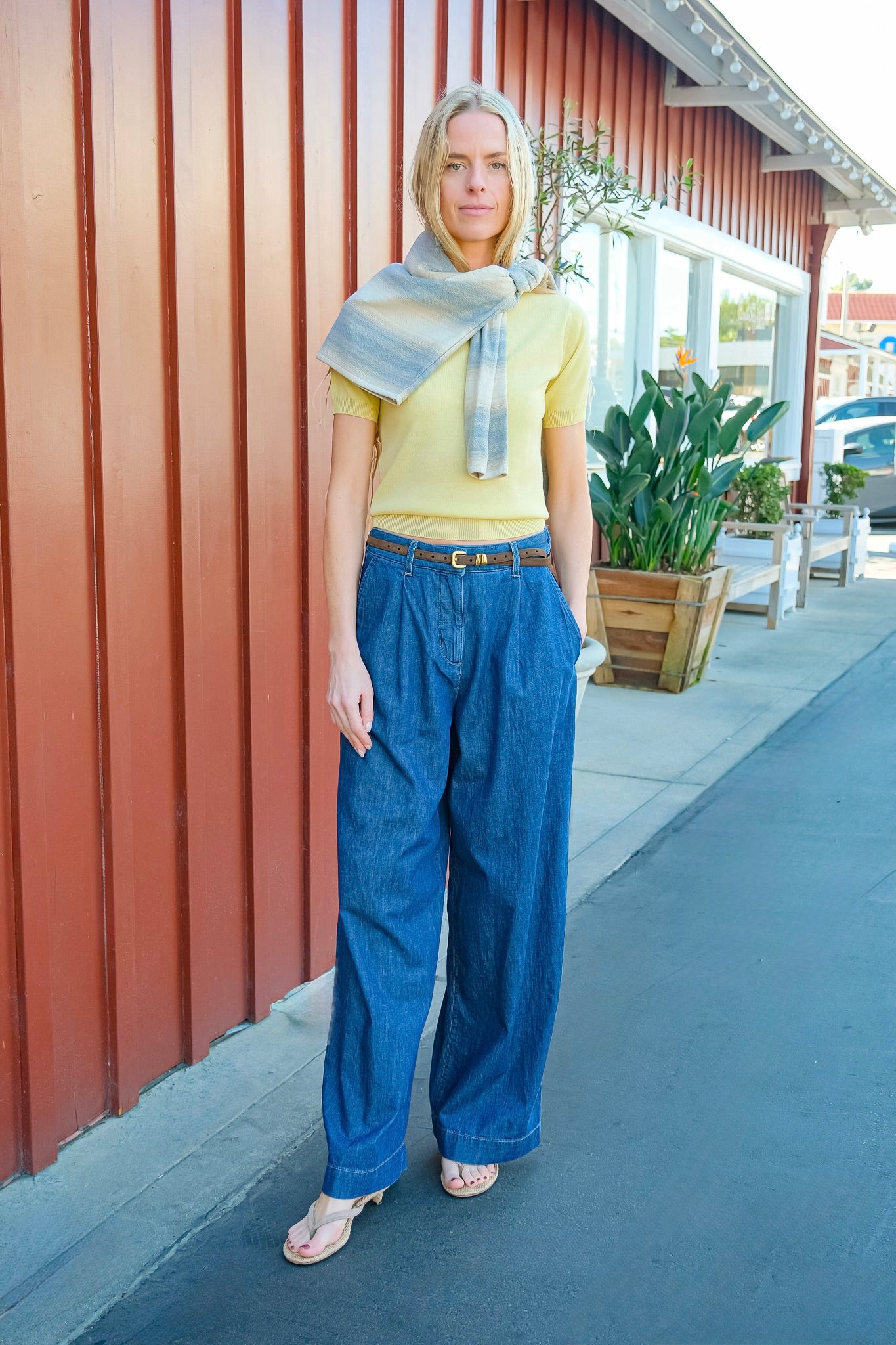 Woman in yellow top and blue jeans standing on a sidewalk with a red building in the background