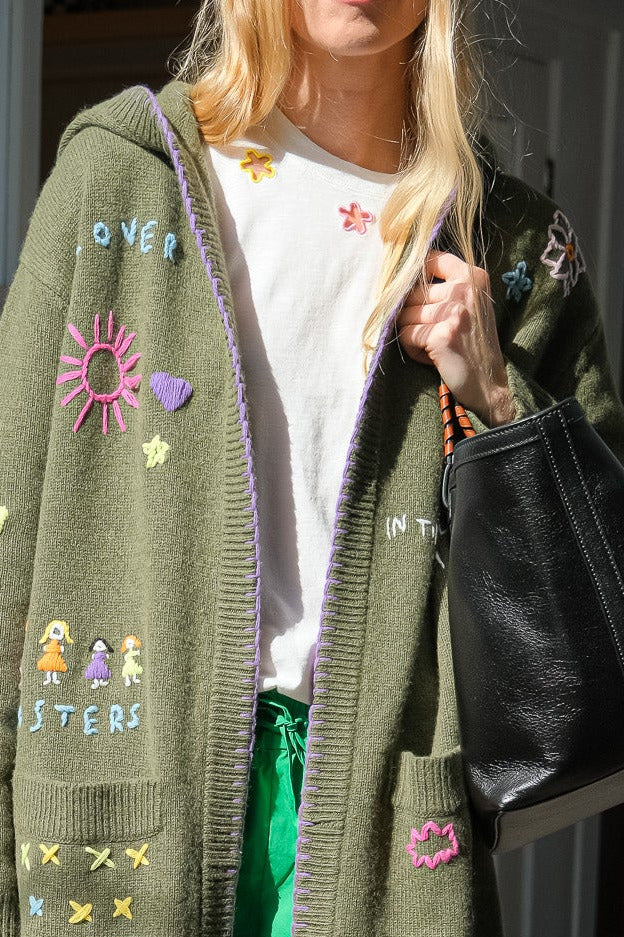 Person wearing a green cardigan with colorful embroidered text and patterns, holding a black bag.