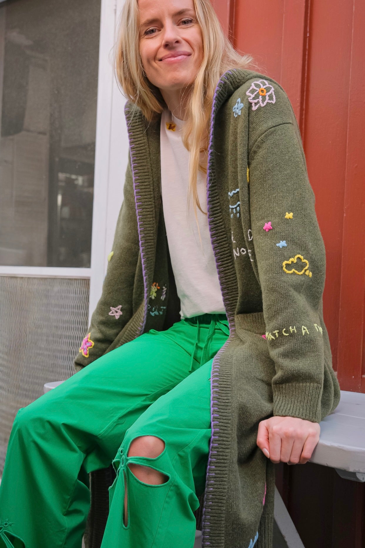 Person wearing a green cardigan with colorful patches and green pants sitting on a bench.