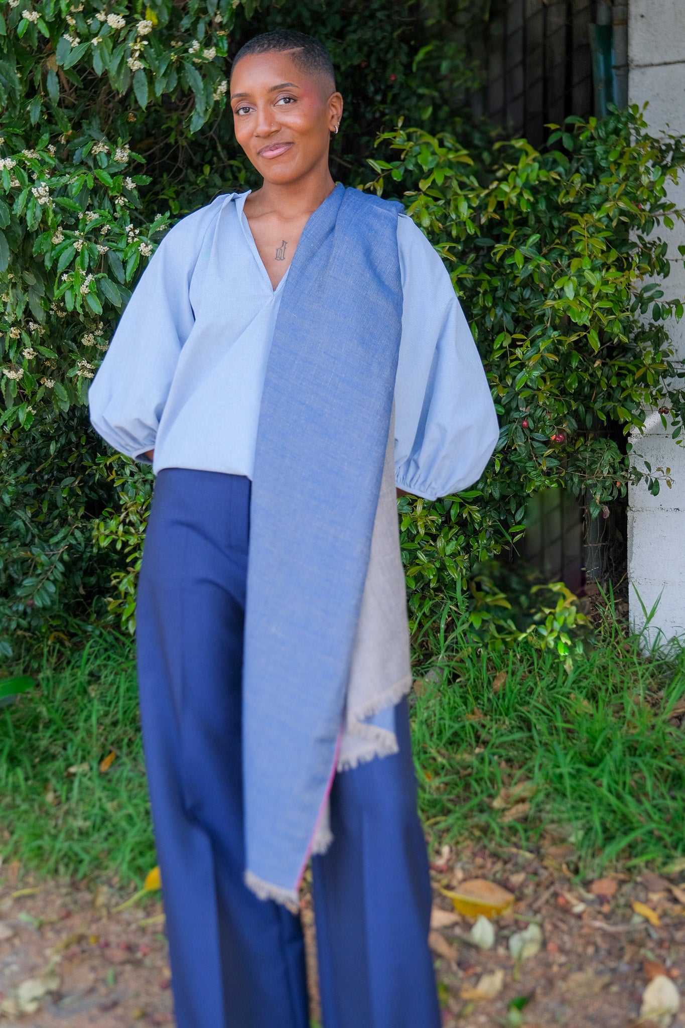 Person wearing a blue scarf and pants standing outdoors with greenery in the background
